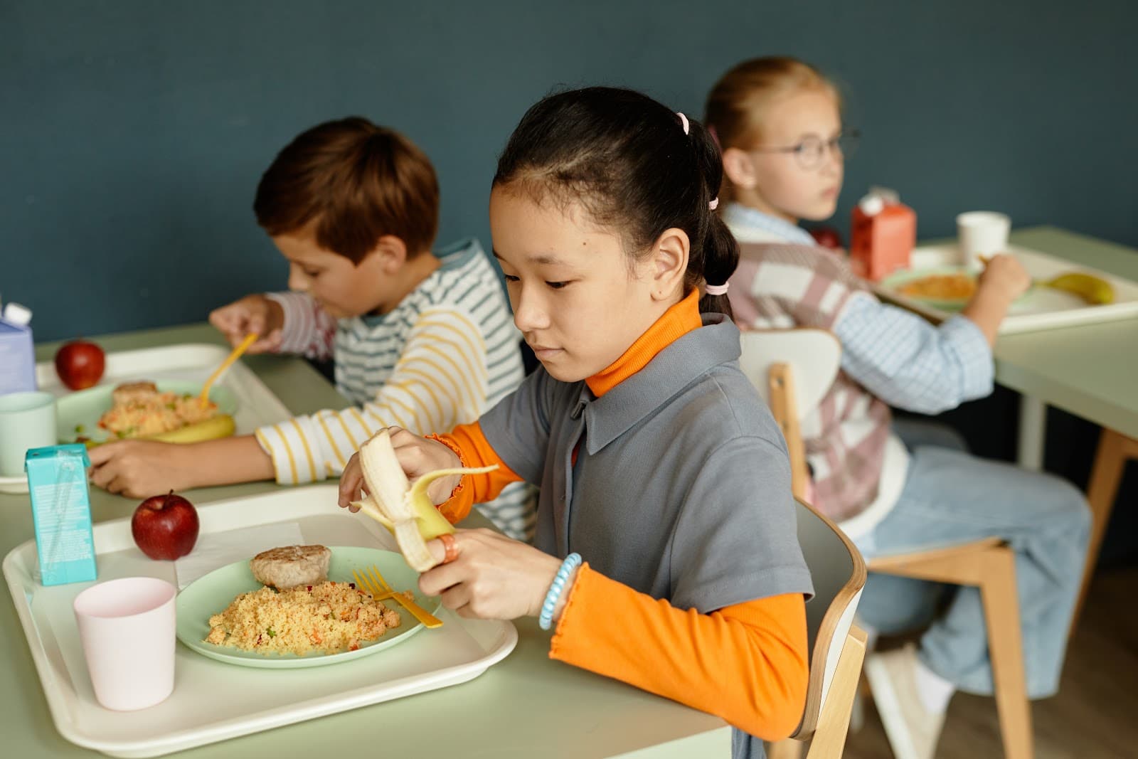 Crianças sentadas à mesa em refeitório escolar interagindo durante a refeição.