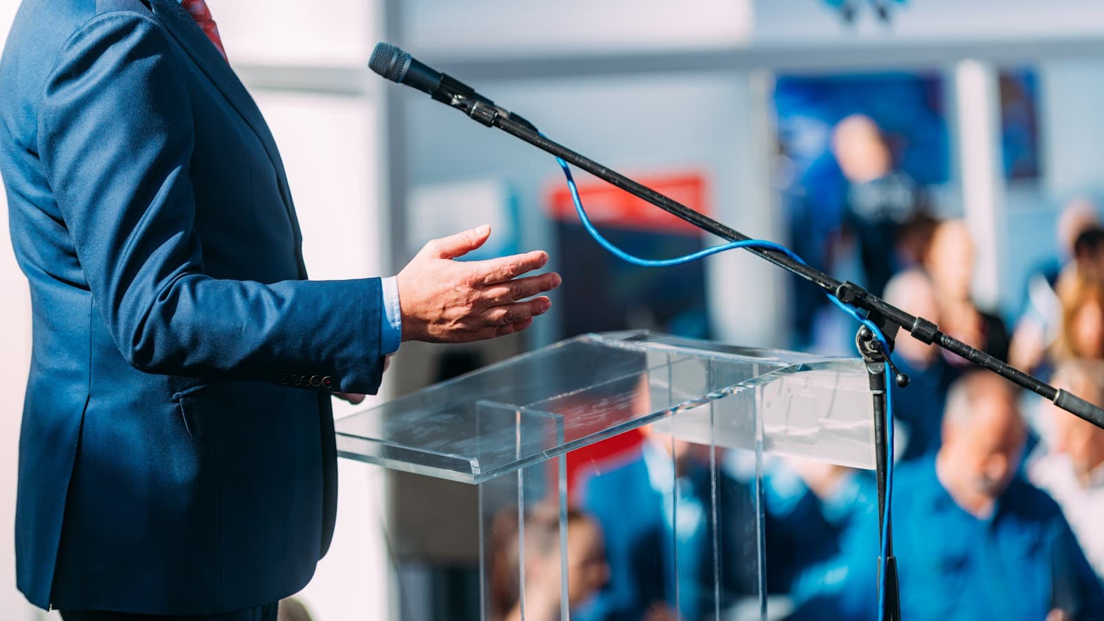 Palestrante profissional discursando em evento com microfone e plateia ao fundo.